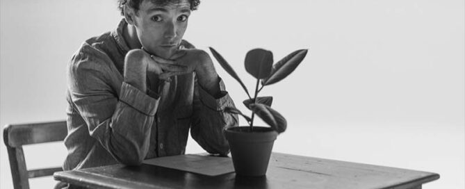 Black and white behind-the-scenes photo of actor Keith Ramsay portraying Francis F. during the filming of the You Were Perfect teaser, seated at a table with a potted plant.
