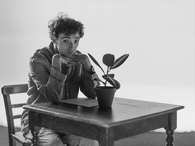 Black and white behind-the-scenes photo of actor Keith Ramsay portraying Francis F. during the filming of the You Were Perfect teaser, seated at a table with a potted plant.