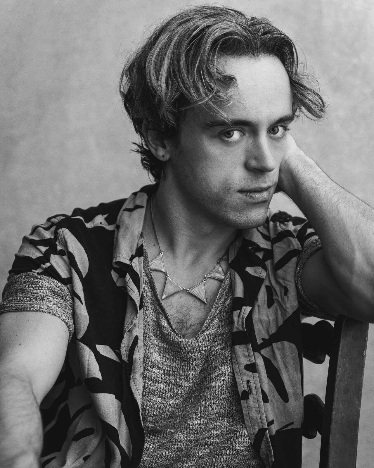 Black and white portrait of actor Bart Lambert seated and gazing at the camera, wearing a patterned shirt and necklace.