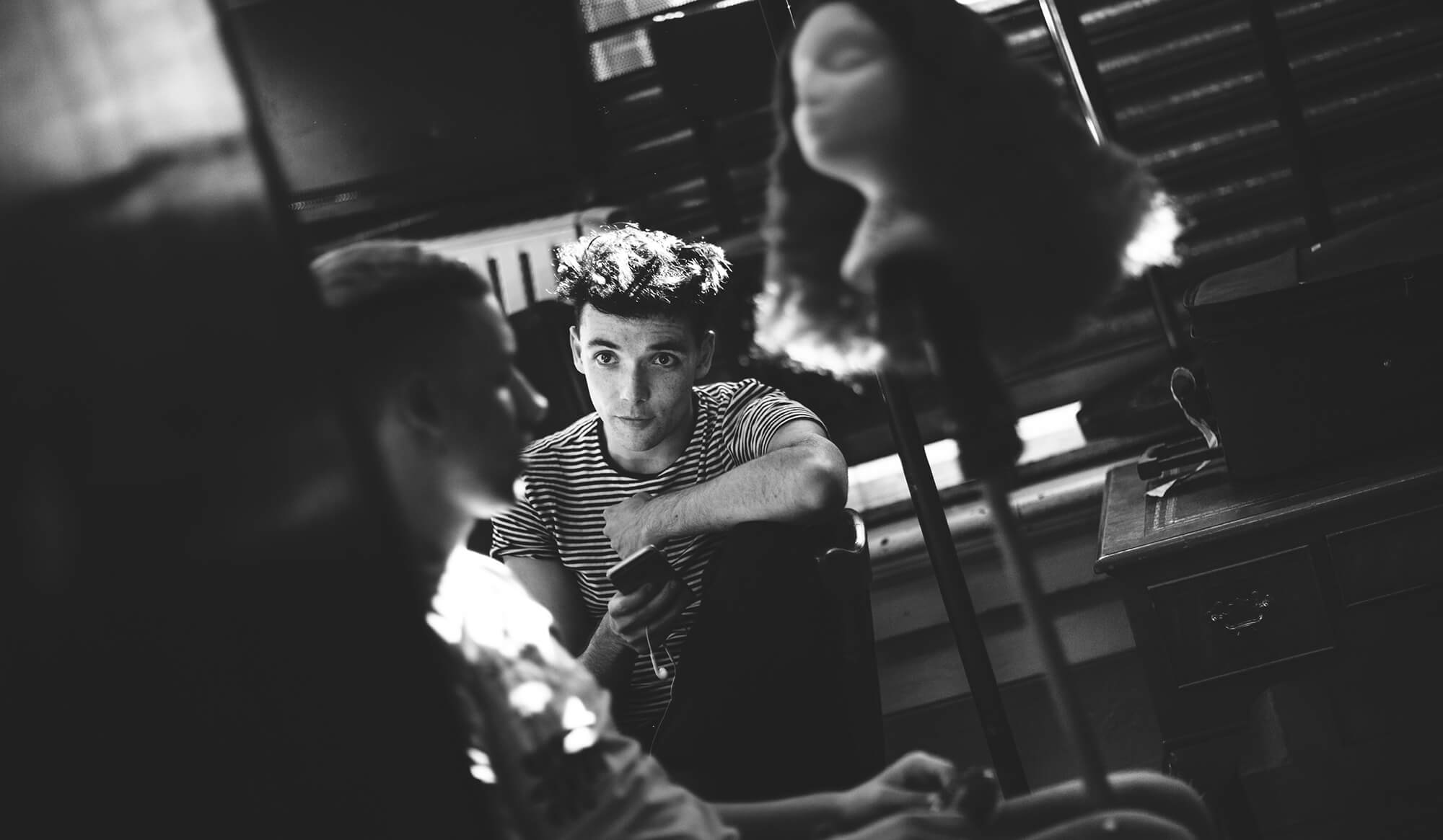 Black and white candid photo of Keith Ramsay seated with a phone in hand, surrounded by props and rehearsal equipment.