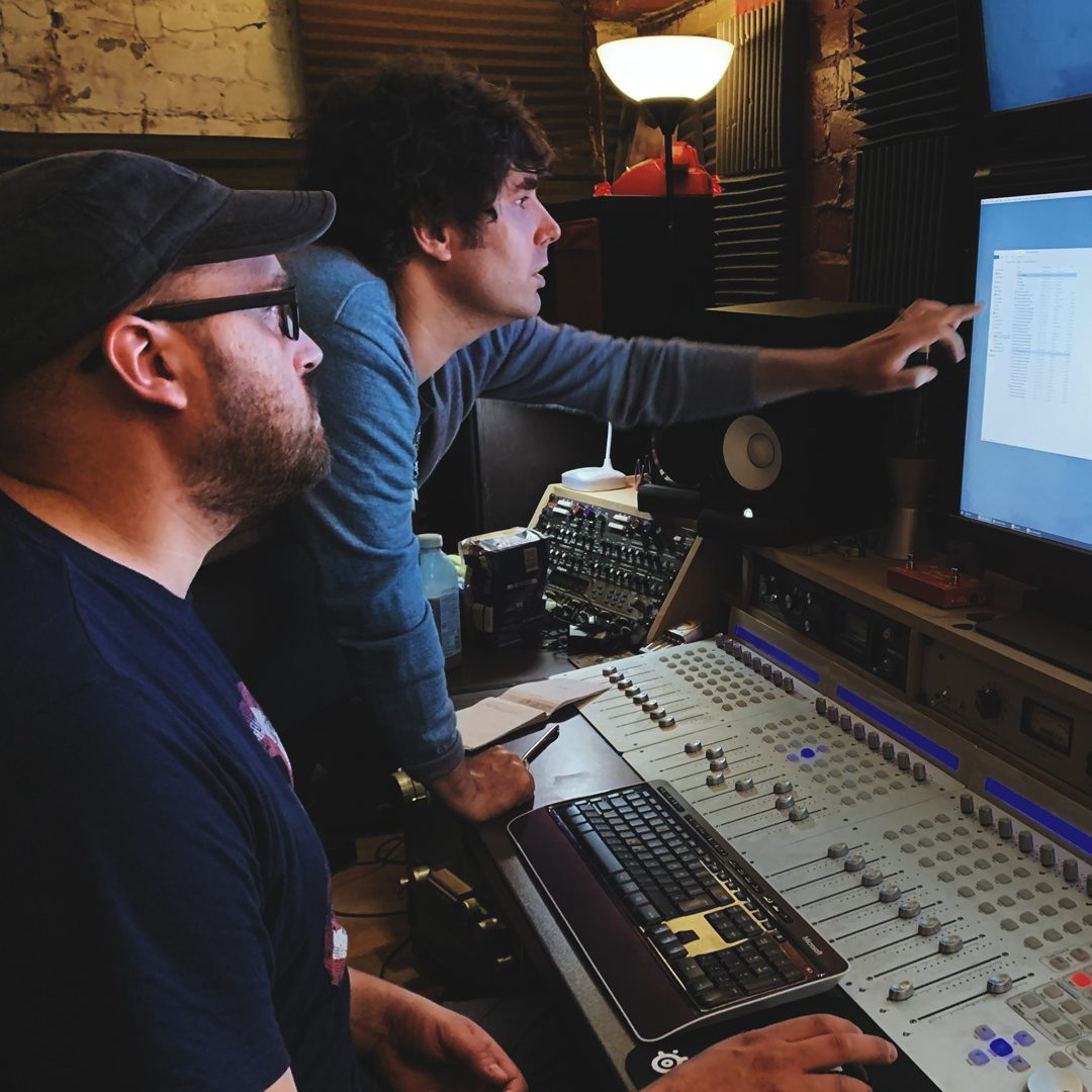 John Wallace, standing and pointing at a monitor in a recording studio, collaborating at a mixing desk with another team member seated beside him.